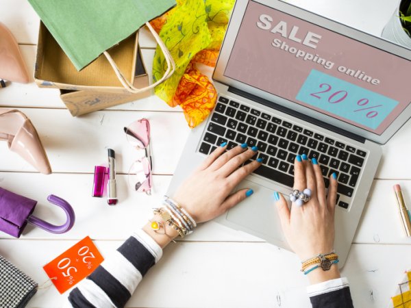 Hände mit bunten Fingernägeln auf der Tastatur eines Laptops, auf dem Display der Schriftzug Online-Shopping, um den Laptop liegen Kleidung und Accessoires