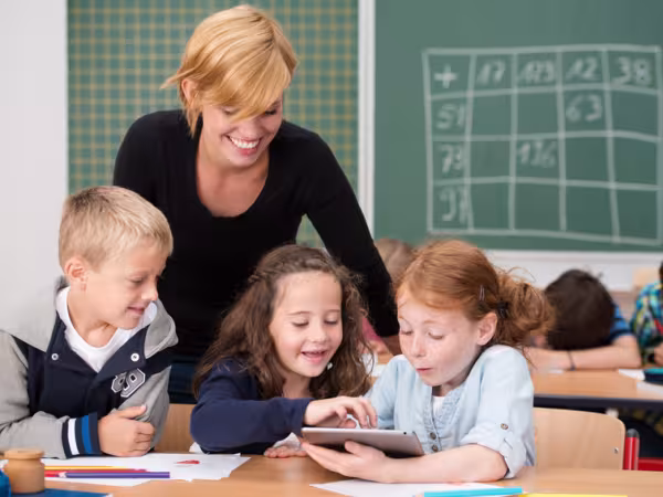 Zu sehen ist eine junge blonde Lehrerin, die an einen Gruppentisch mit drei kleinen Schulkindern steht. Sie beugt sich hinab zu dem Jungen und den beiden Mädchen, die gemeinsam an einem Tablett arbeiten. Im Hintergrund sind weitere Schulkinder und eine Tafel zu sehen. 