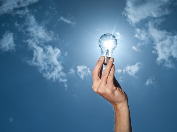 Zu sehen ist ein ausgestreckter Arm vor blauem Himmel. Die Finger umklammern das Gewinde einer gäsernen Glühbirne. Die strahlende Sonne scheint den Glühdraht zum Strahlen zu bringen. 