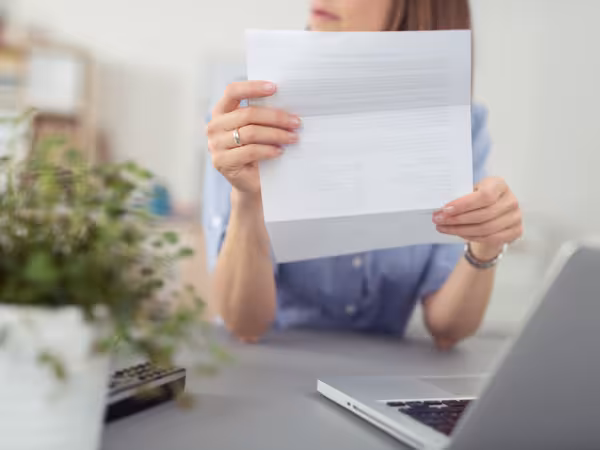 Eine Person sitzt an einem Tisch vor einem Laptop und hält einen Brief hoch.
