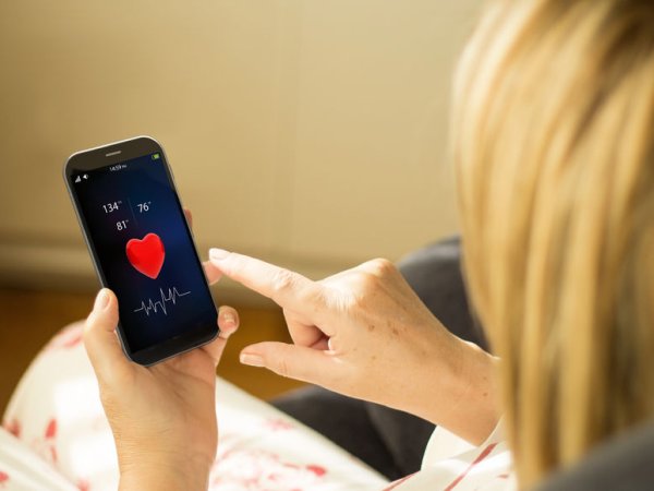 Zu sehen ist der Kopf einer blonden Frau. Sie hält ein Smartphone in den Händen, auf dem ein rotes Herz und Angaben zur Gesundheit zu sehen sind. 