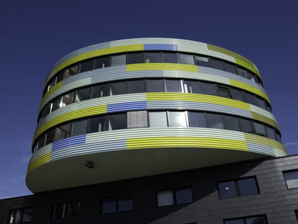 Zu sehen ist ein ellipsenförmiges dreistöckiges Gebäudes mit umlaufenden Fenstern. Die Fassadengestaltung ist neongelb und hebt sich um blauen Himmel ab. Bei der sogenannte Pillbox handelt es sich um ein Gebäudeteil, in dem der vzbv seine Büroräume hat. 