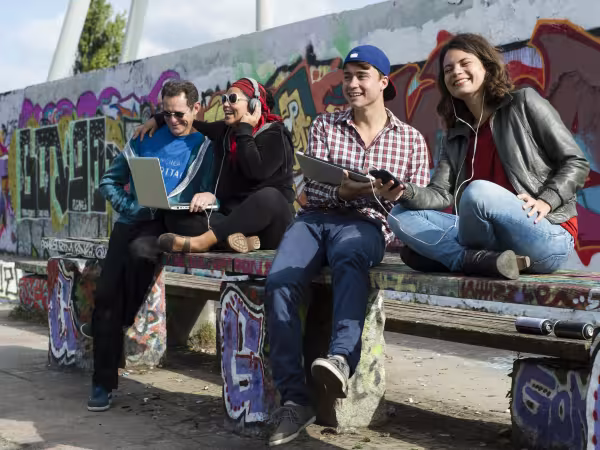 Zu sehen sind vier junge Leute vor einer Wand mit Graffiti. Paarweise sind die Männer und Frauen mit Laptop und Kopfhörern ausgestattet. Sie schauen in die Ferne bzw. in die Geräte und sind positiver Stimmung. 