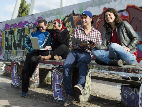 Zu sehen sind vier junge Leute vor einer Wand mit Graffiti. Paarweise sind die Männer und Frauen mit Laptop und Kopfhörern ausgestattet. Sie schauen in die Ferne bzw. in die Geräte und sind positiver Stimmung.