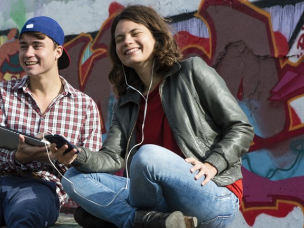 Zu sehen sind vier junge Leute vor einer Wand mit Graffiti. Paarweise sind die Männer und Frauen mit Laptop und Kopfhörern ausgestattet. Sie schauen in die Ferne bzw. in die Geräte und sind positiver Stimmung.