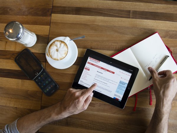 You can see a wooden table with a cup of Coffee an a phone, an open notebook and a tablet. The website of the conxumer advice center ist open on the tablet. 